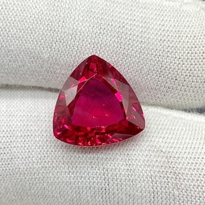 Op de afbeelding: Een driehoekige, gefacetteerde edelsteen met een dieprode kleur. De edelsteen is geplaatst tegen een witte achtergrond.
