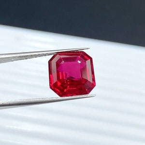May include: A deep red gemstone with a square cut, held by tweezers against a white background.
