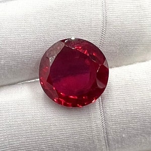 May include: A deep red gemstone with a round, faceted cut. The gemstone is set against a white background.