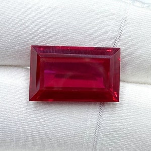 Può includere: Una gemma rettangolare, sfaccettata, di colore rosso intenso. La gemma è posta su uno sfondo bianco, mostrando la sua chiarezza e il suo colore. Il taglio della pietra è a forma di baguette, con linee pulite e dritte e una superficie lucida. La gemma è probabilmente un rubino.