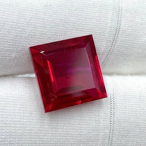 Könnte beinhalten: Ein leuchtend roter quadratischer Edelstein mit facettiertem Schliff. Der Edelstein wird auf einem weißen Stoffhintergrund präsentiert.