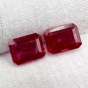 May include: Two rectangular-cut, deep red gemstones. The gems are displayed on a white, textured surface. The stones have a polished, reflective surface, showcasing their clarity and rich color. The gems are likely rubies, perfect for jewelry.