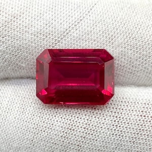 May include: A deep red gemstone with a rectangular emerald cut. The gemstone is displayed on a white fabric background.