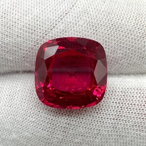 May include: A deep red gemstone with a cushion cut. The gemstone is set on a white fabric background.