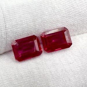 Puede incluir: Dos piedras preciosas de color rojo talla esmeralda sobre un fondo de tela blanca.