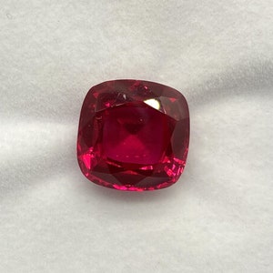 Puede incluir: Una piedra preciosa de color rojo intenso con talla cojín, expuesta en una caja forrada de terciopelo blanco.