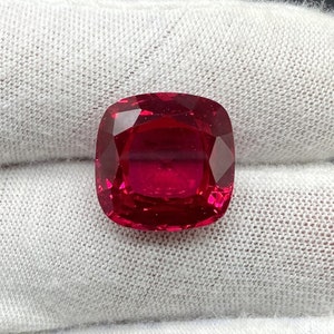 Może przedstawiać: Jaskrawoczerwony kamień szlachetny o szlifie poduszkowym. Kamień szlachetny jest umieszczony na białym tle z tkaniny.