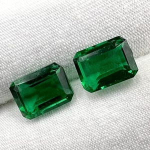 May include: Two emerald green gemstones, cut in a rectangular emerald shape, are displayed on a white fabric background.