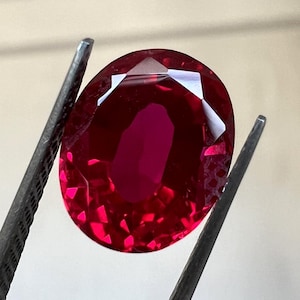 May include: A deep red oval-shaped gemstone held by tweezers. The gemstone is faceted and has a high polish.