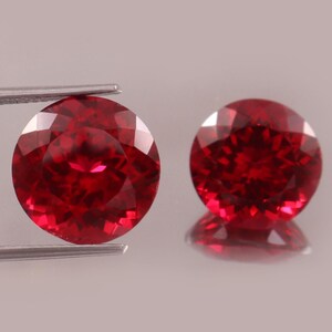 Op de afbeelding: Twee ronde, gefacetteerde, dieprode edelstenen. Eén wordt vastgehouden door een zilveren pincet, de andere ligt op een reflecterend oppervlak. De edelstenen hebben een briljante slijpvorm, die hun interne structuur en kleur benadrukt.