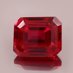 May include: A faceted, rectangular-cut ruby gemstone. The gem is a deep, saturated red color with a high degree of clarity. The cut of the stone creates multiple facets that reflect light, giving it a brilliant appearance. The ruby is set against a neutral background.