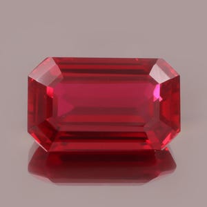 Könnte beinhalten: Ein facettierter, rechteckig geschliffener Rubin-Edelstein. Der Edelstein hat eine tiefe, leuchtende rote Farbe mit einer reflektierenden Oberfläche. Der Smaragdschliff weist eine Reihe paralleler Facetten auf, die ein brillantes Spiel von Licht und Farbe erzeugen. Der Edelstein ist vor einem neutralen Hintergrund platziert.