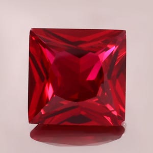 May include: A square-cut, faceted ruby gemstone. The gem is a deep, vibrant red color, with sharp, clean lines and a polished surface. The gemstone is set against a soft, neutral background, highlighting its brilliance.