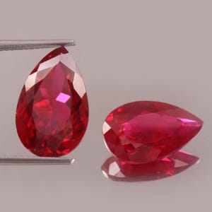 Puede incluir: Dos rubíes facetados en forma de pera. Uno está sujeto con pinzas, el otro descansa sobre una superficie reflectante. Las gemas son de un rojo intenso y vibrante, con múltiples facetas que captan la luz. El tema general son las piedras preciosas.