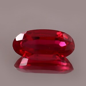 May include: A vibrant, oval-cut ruby gemstone. The gem is a deep, saturated red with facets that catch the light, creating brilliant reflections. The ruby is displayed on a reflective surface, enhancing its color and shape.