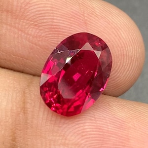 Könnte beinhalten: Ein facettierter, ovalförmiger, tiefroter Edelstein. Der Edelstein wird in einer Hand vor einem grauen Hintergrund gehalten. Die Facetten des Steins reflektieren das Licht und zeigen seine Brillanz und Farbe. Dieser Edelstein könnte in Schmuck verwendet werden.