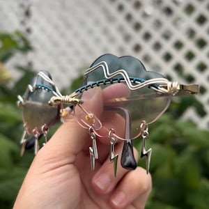 May include: Cloud-shaped sunglasses with pink lenses and silver lightning bolt charms hanging from the bottom. The sunglasses are made of wire and have a blue and silver design.