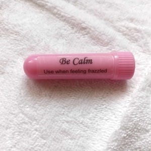 May include: A pink tube with the words "Be Calm" and "Use when feeling frazzled" printed on it. The tube has a rounded end and a ridged cap. The product is laying on a white, textured surface.