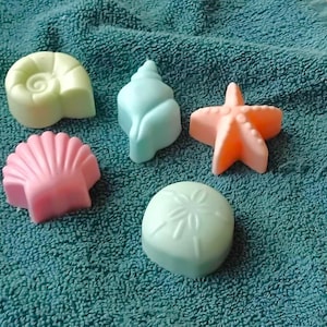 May include: Five handmade soap bars shaped like seashells and a starfish. The soaps are in shades of green, blue, pink, and orange. The soaps are on a teal towel.