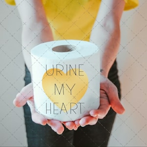 Puede incluir: Una persona sosteniendo un rollo de papel higiénico blanco con el texto "URINE MY HEART" y un corazón amarillo.