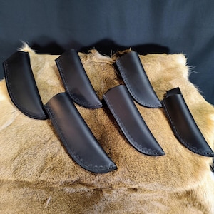 May include: Six black leather knife sheaths with a decorative stitch around the edges, displayed on a brown fur surface.