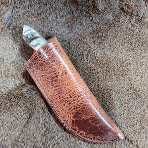 May include: A brown leather knife sheath with a decorative stitching detail. The sheath is lying on a brown fur surface.