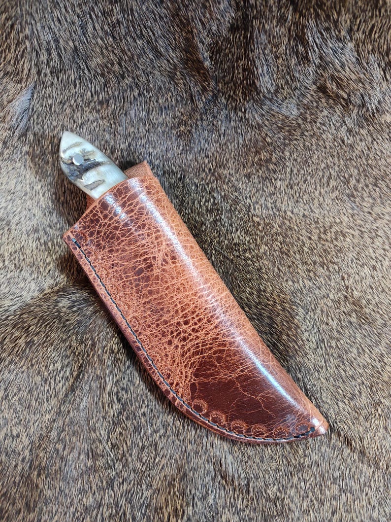 May include: A brown leather knife sheath with a decorative stitching detail. The sheath is lying on a brown fur surface.