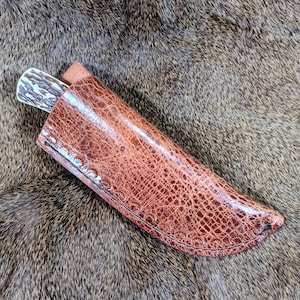 May include: A brown leather knife sheath with a stitched edge. The sheath has a textured, alligator-like pattern.