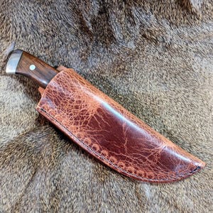 May include: A brown leather knife sheath with a stitched edge. The sheath is lying on a brown fur surface.