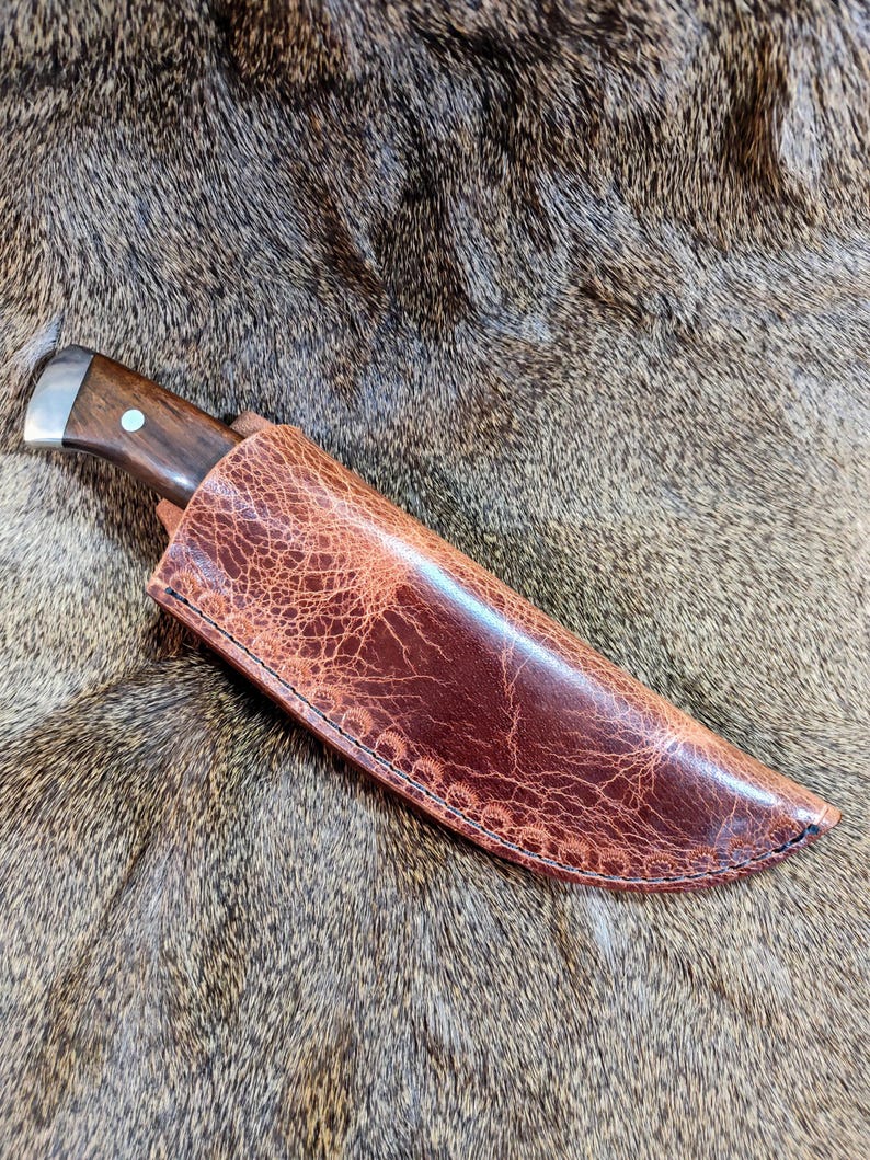 May include: A brown leather knife sheath with a stitched edge. The sheath is lying on a brown fur surface.