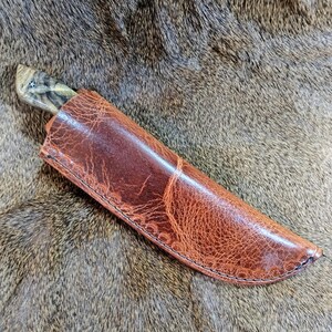 May include: A brown leather knife sheath with a stitched edge. The sheath is lying on a brown fur surface.