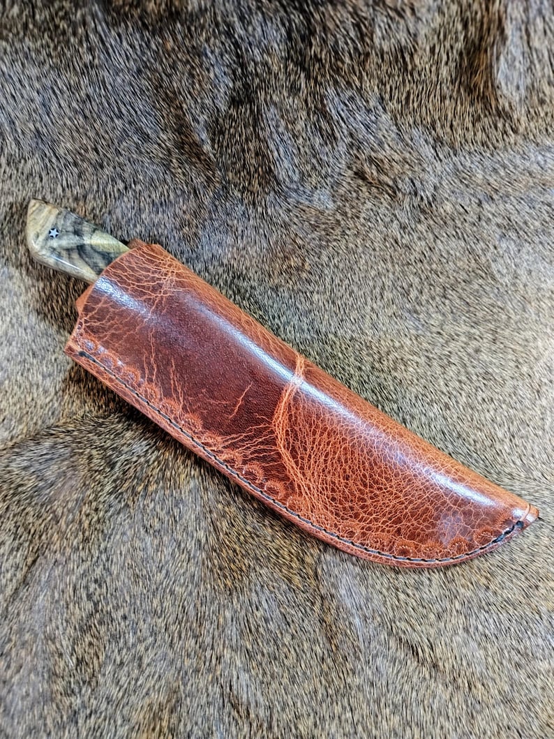 May include: A brown leather knife sheath with a stitched edge. The sheath is lying on a brown fur surface.