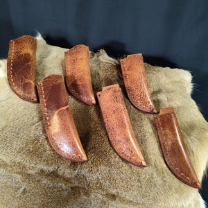 May include: Six brown leather knife sheaths with a textured surface. The sheaths are arranged on a light brown fur surface.