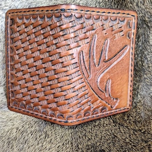 May include: A brown leather wallet with a woven pattern and a tooled design of an antler. The wallet has a black stitching detail around the edges.