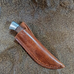 May include: A brown leather knife sheath with a stitched edge. The sheath is lying on a brown fur surface.
