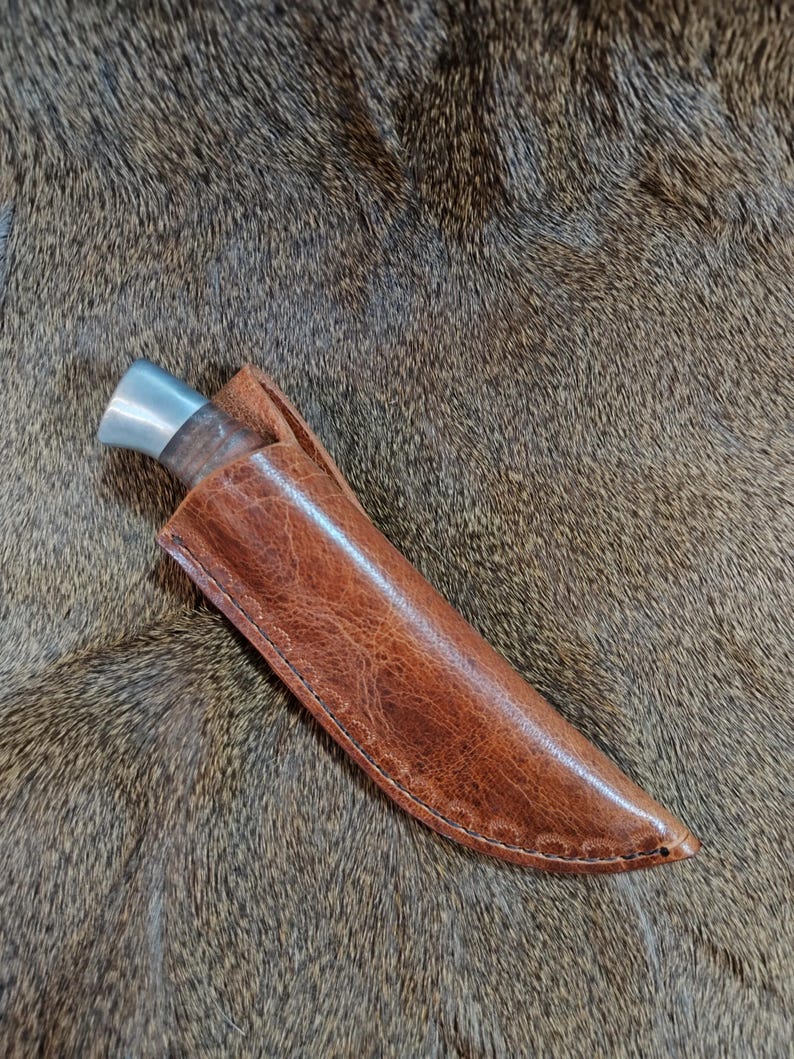 May include: A brown leather knife sheath with a stitched edge. The sheath is lying on a brown fur surface.