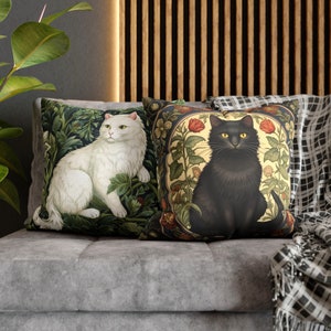 May include: Two decorative throw pillows with cat illustrations. One pillow features a white cat amidst green foliage. The other pillow showcases a black cat surrounded by red roses and floral patterns. Both pillows are on a gray sofa.