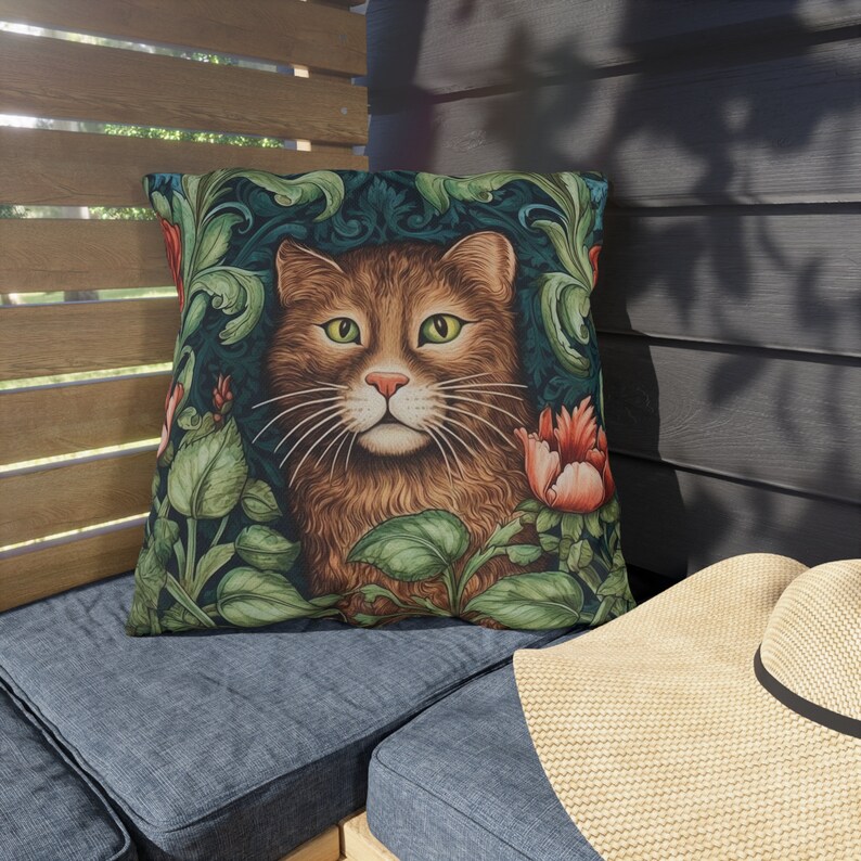 May include: Square throw pillow featuring a detailed illustration of a brown cat with green eyes, surrounded by green foliage and red flowers. The pillow has a dark teal background and is placed on a blue cushion.