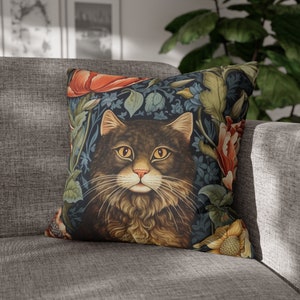 May include: Square throw pillow featuring a detailed illustration of a fluffy brown cat with yellow eyes, surrounded by a floral pattern of red, yellow, and green flowers and leaves on a dark blue background. The pillow is placed on a gray sofa.