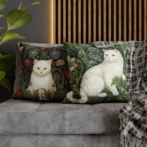 May include: Two decorative throw pillows featuring white cats amidst a dark green floral pattern. The cats have yellow eyes and are surrounded by detailed foliage. The pillows are placed on a gray sofa, creating a cozy and artistic home decor.