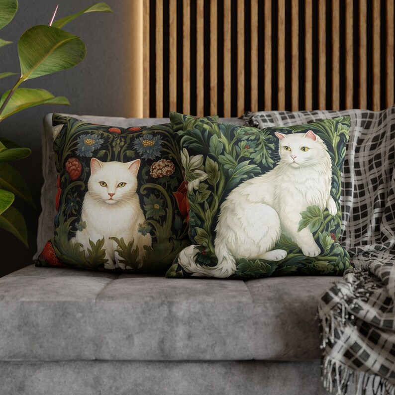 May include: Two decorative throw pillows featuring white cats amidst a dark green floral pattern. The cats have yellow eyes and are surrounded by detailed foliage. The pillows are placed on a gray sofa, creating a cozy and artistic home decor.