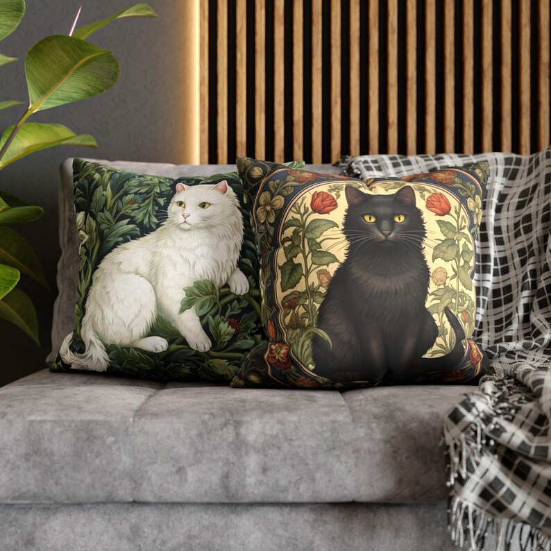 May include: Two decorative throw pillows with cat illustrations. One pillow features a white cat amidst green foliage. The other pillow showcases a black cat surrounded by red roses and floral patterns. Both pillows are on a gray sofa.