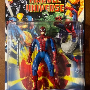 Op de afbeelding: Een rood en blauw action figure van Spider-Man met een gescheurd kostuum. De figuur houdt een web shooter in de ene hand en heeft een rugzak op zijn rug. De verpakking heeft een achtergrond met andere Marvel-personages, waaronder Hulk en Thing. De tekst op de verpakking luidt "Battle Ravaged Spider-Man Secret Storage Backpack".