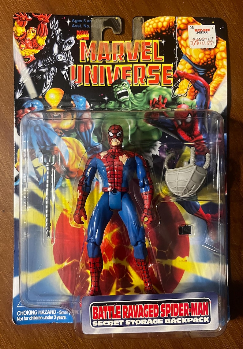 Op de afbeelding: Een rood en blauw action figure van Spider-Man met een gescheurd kostuum. De figuur houdt een web shooter in de ene hand en heeft een rugzak op zijn rug. De verpakking heeft een achtergrond met andere Marvel-personages, waaronder Hulk en Thing. De tekst op de verpakking luidt "Battle Ravaged Spider-Man Secret Storage Backpack".