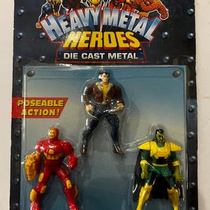 May include: A sealed package of "Heavy Metal Heroes" die-cast metal action figures. The package features three figures: Iron Man in red and gold, a figure in brown and blue, and a green and yellow figure. The package has the Marvel Comics logo.