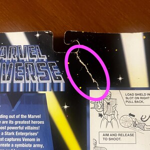 Pode incluir: Um close-up de uma embalagem de figura de a&ccedil;&atilde;o do Marvel Universe com uma rachadura no meio. A embalagem apresenta uma ilustra&ccedil;&atilde;o em preto e branco da figura de a&ccedil;&atilde;o com um lan&ccedil;ador de teias e o texto "LOAD SHIELD INTO SLOT ON RIGHT ARM... PULL BACK." e "AIM AND RELEASE TO SHOOT."
