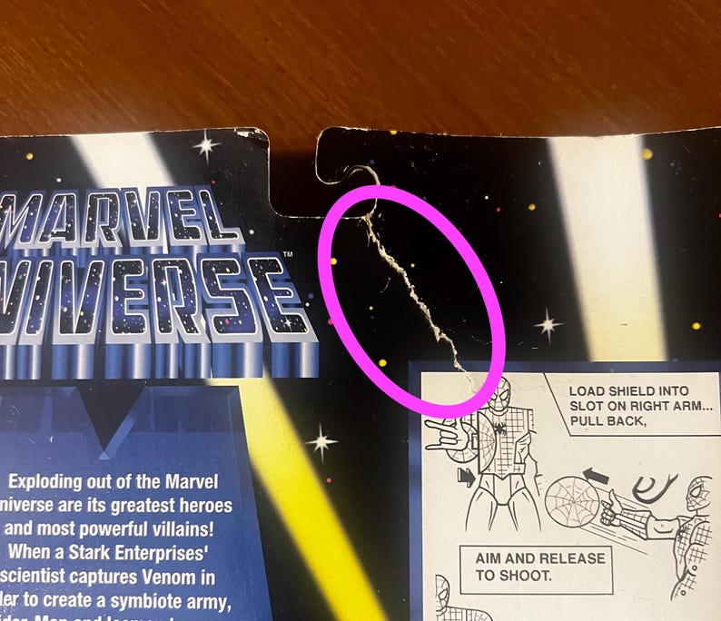 Pode incluir: Um close-up de uma embalagem de figura de a&ccedil;&atilde;o do Marvel Universe com uma rachadura no meio. A embalagem apresenta uma ilustra&ccedil;&atilde;o em preto e branco da figura de a&ccedil;&atilde;o com um lan&ccedil;ador de teias e o texto "LOAD SHIELD INTO SLOT ON RIGHT ARM... PULL BACK." e "AIM AND RELEASE TO SHOOT."