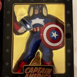 May include: Captain America action figure in a display box. The figure is in a blue and white costume, holding a shield with a white star and red and white stripes. The box has the words "Captain America" in a bold font.