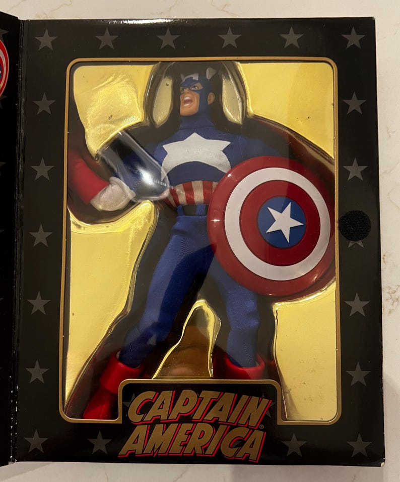 May include: Captain America action figure in a display box. The figure is in a blue and white costume, holding a shield with a white star and red and white stripes. The box has the words "Captain America" in a bold font.
