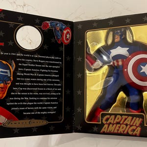 May include: A Captain America action figure in a display box. The figure is in a blue, red, and white costume, holding a shield. The box has a gold interior and black exterior with text that reads "Captain America."